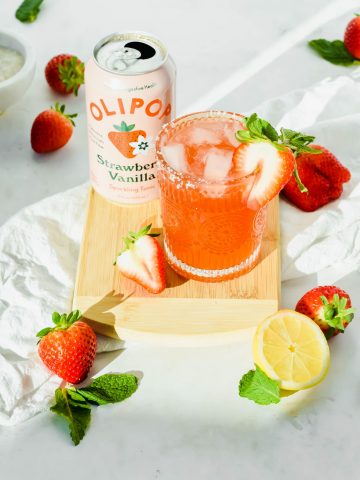 Strawberry Mint Sunshine Cocktail and a can of OLIPOP Strawberry Vanilla on a wooden cutting board surrounded my fresh strawberries, mint leaves, and half a lemon