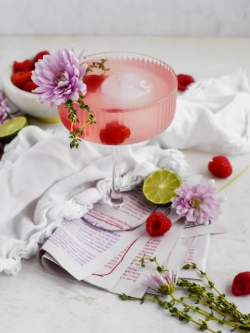 Raspberry Hibiscus Cocktail in a stemmed cocktail glass with ice garnished with fresh flowers, thyme, and raspberries