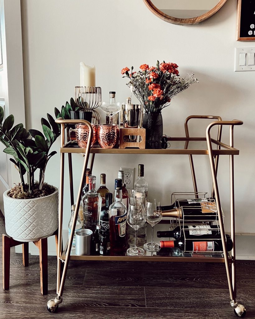 full stocked bar cart with alcohol, glasses, mugs, plants beside a plant in a white vase against white wall