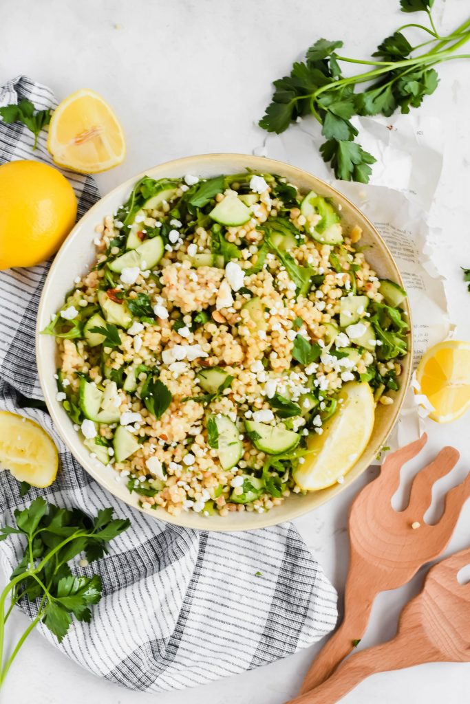 Overhead shot of Lemon Arugula Couscous Salad in a big serving bowl surrounded by fresh lemons and herbs