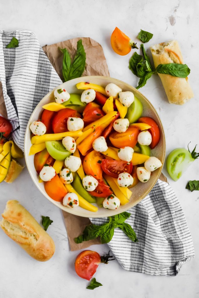 Overhead view of Mango Caprese Salad in a large round bowl surrounded by fresh bread, tomato slices, sliced mango, basil leaves, and a slotted spoon
