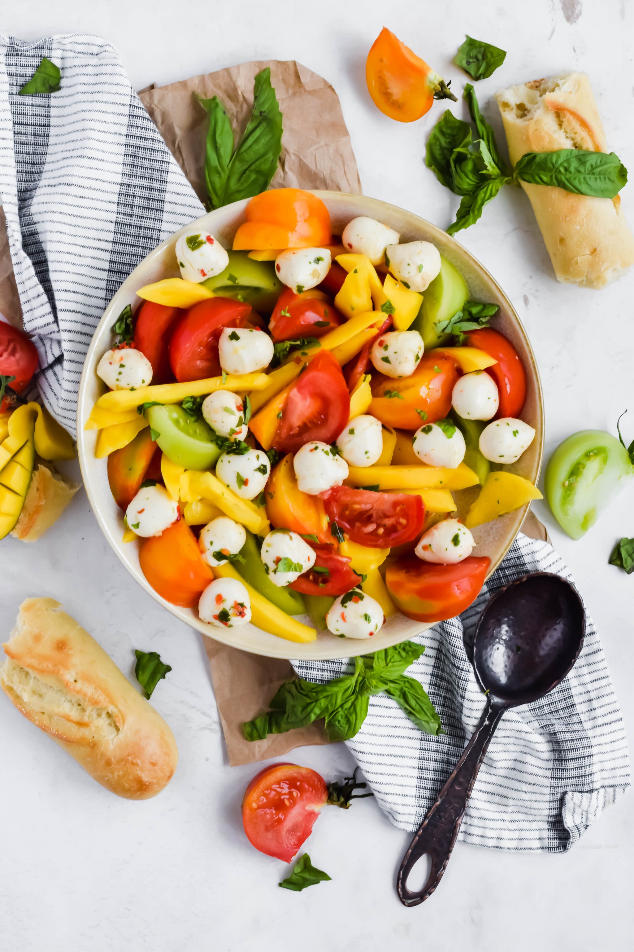 Mango tomato Salad in a large round bowl surrounded by bread, tomatoes, mango, basil, and a slotted spoon.