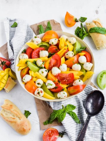 Overhead view of Mango Caprese Salad in a large round bowl surrounded by fresh bread, tomato slices, sliced mango, basil leaves, and a slotted spoon