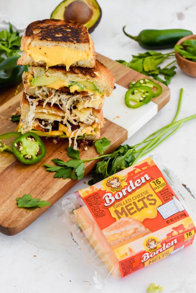 four jalapeño popper grilled cheese halves stacked on top of eachother on wood board surrounded by fresh herbs and jalapeño beside a package of Borden cheese melts