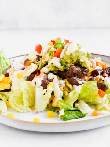 lettuce, fresh veggies, and steak with scoops of guacamole on top drizzled with creamy dressing.