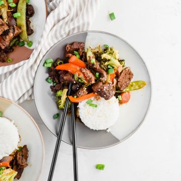 plated beef and broccoli stir fry beside bed of white rice on gray plate garnished with green onion on white background with two additional servings plated and running out of photo frame.