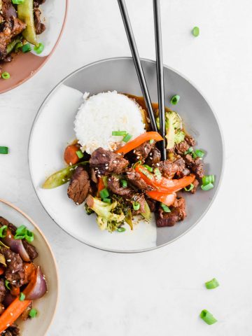 plated beef and broccoli stir fry beside bed of white rice on gray plate garnished with green onion and chop stick on the plate on white background.