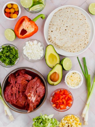 Ingredients for Carne Asada Street Beef Tacos including raw steak, avocado, diced bell pepper, diced onion, corn, shredded lettuce, green onions, coquito cheese, cilantro, cherry tomatoes, limes, and tortillas
