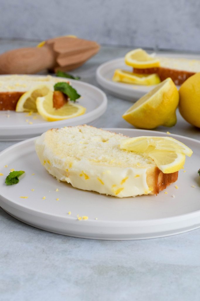 side view of slice of glazed lemon bread garnished with fresh lemon slice on white plate with additional bread slices and lemon in background.