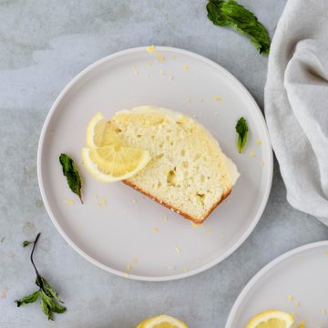 overhead shot of one slice of lemon quick bread on white plate garnished with lemon slices and mint leaves on gray background.