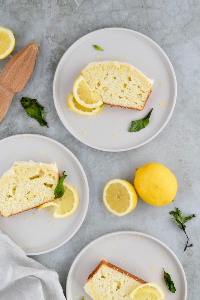 overhead shot of three slices of lemon quick bread on white plates garnished with lemon slices and mint leaves on gray background.