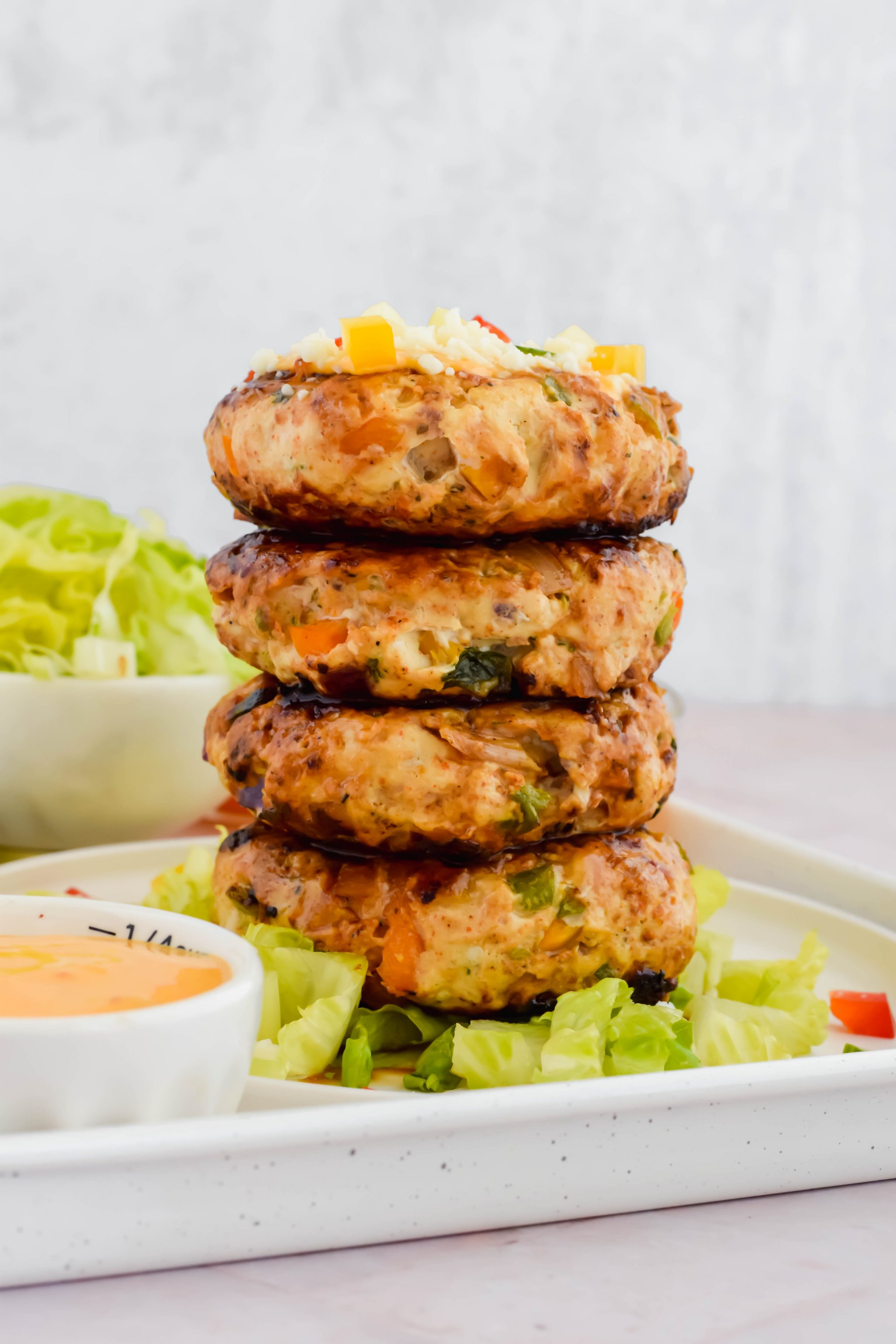 4 cooked fajita burgers stacked on top of one another on top of bed of lettuce beside bowl of orange sauce.