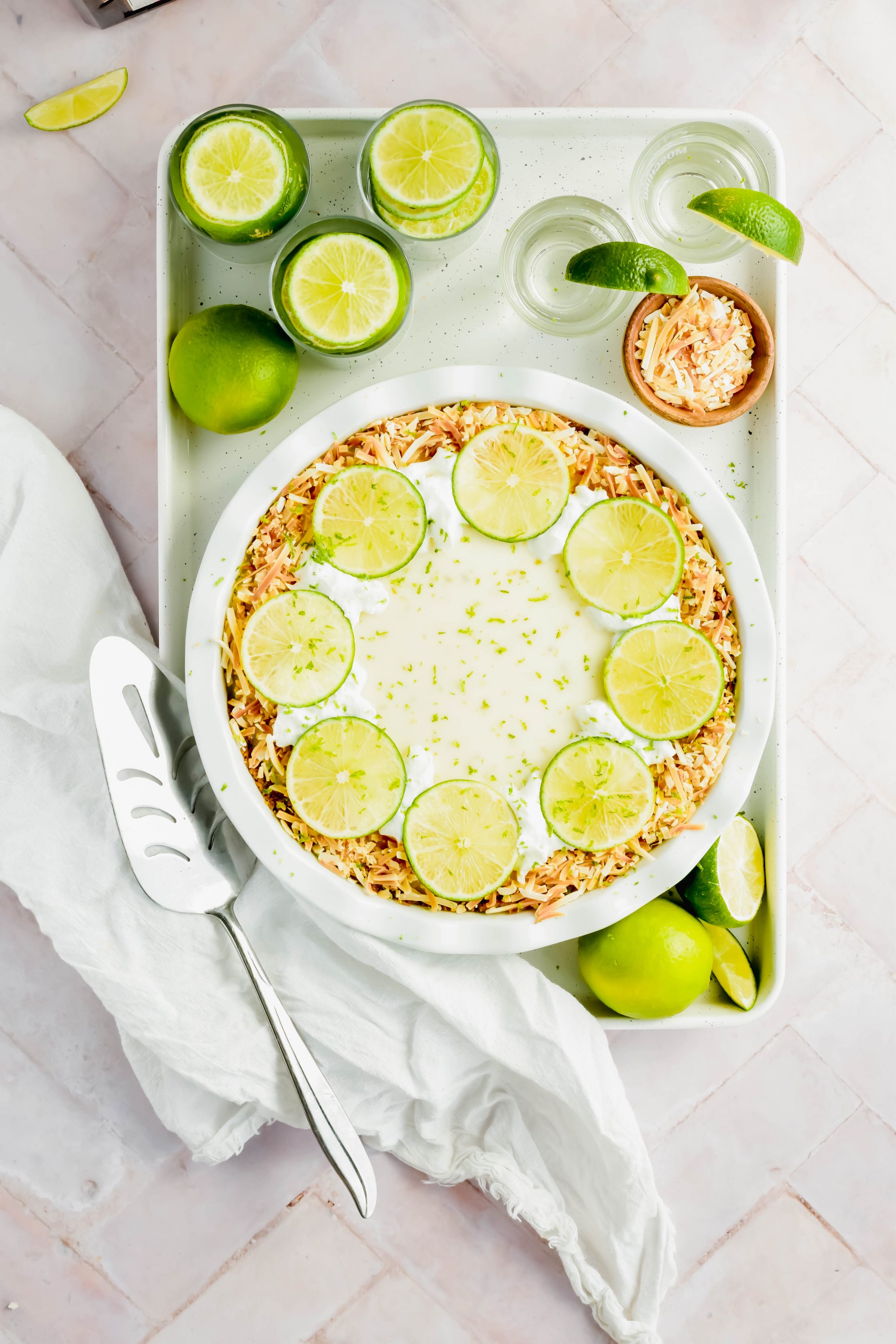 key lime pie garnished with toasted coconut and lime slices on white baking sheet surrounded by limes and tequila shots on white background.
