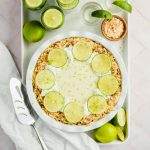 key lime pie garnished with toasted coconut and lime slices on white baking sheet surrounded by limes and tequila shots with white background.