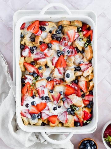 overhead shot of baked healthy french toast casserole garnished with fresh blueberries and strawberries with white glaze drizzled over top in white baking tray on white background..