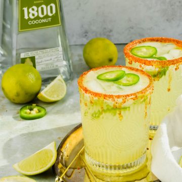 Two Spicy Skinny Jalapeno Margaritas on a gold serving tray next to toothpicks, limes, and a bottle of 1800 Coconut tequila