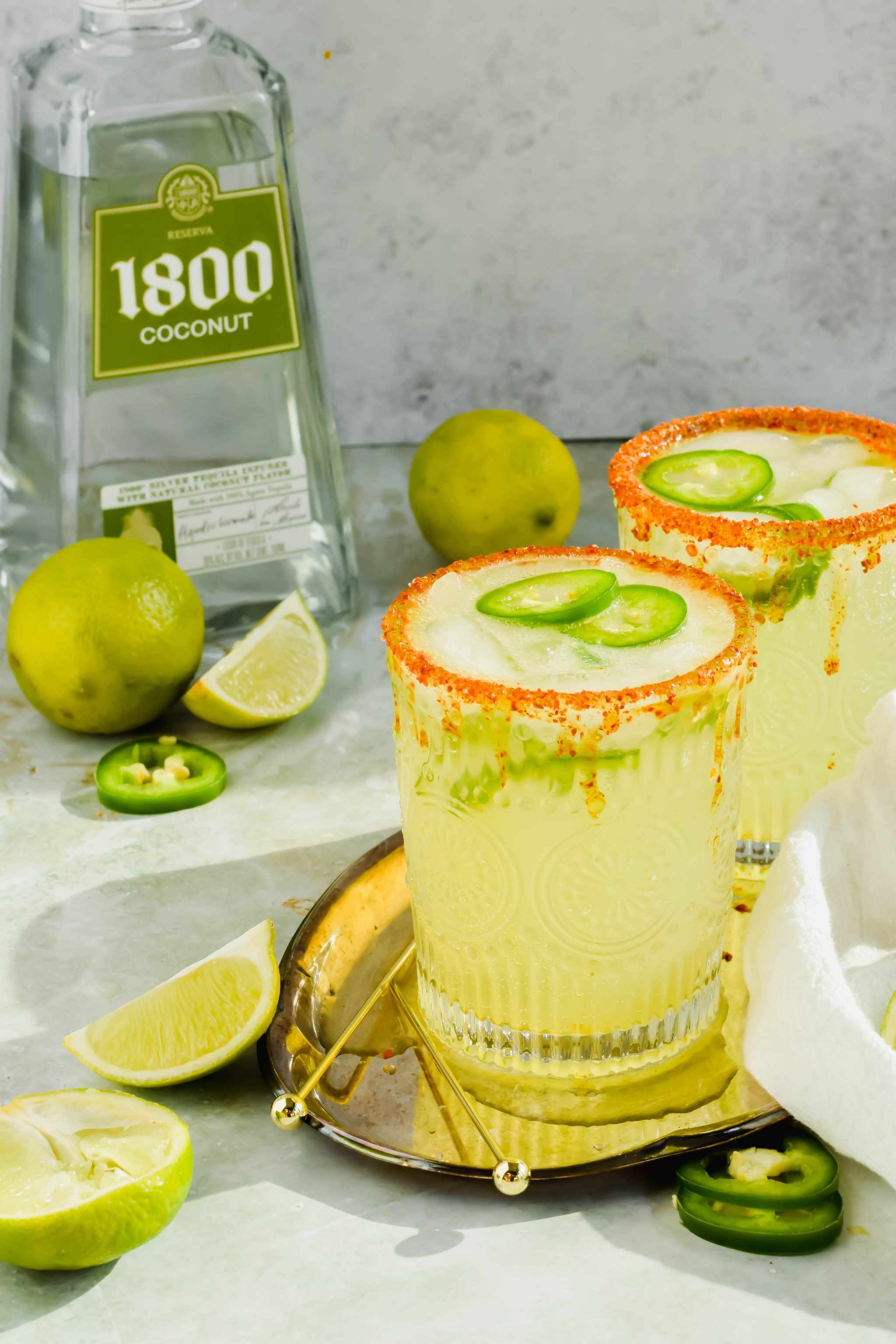 Two Spicy Skinny Jalapeno Margaritas on a gold serving tray next to toothpicks, limes, and a bottle of 1800 Coconut tequila