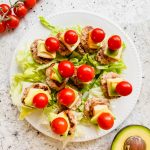 lettuce underneath a mini turkey burger on a stick topped with cheese, avocado and a cherry tomato on white plate on marble counter beside additional tomatoes and half an avocado..
