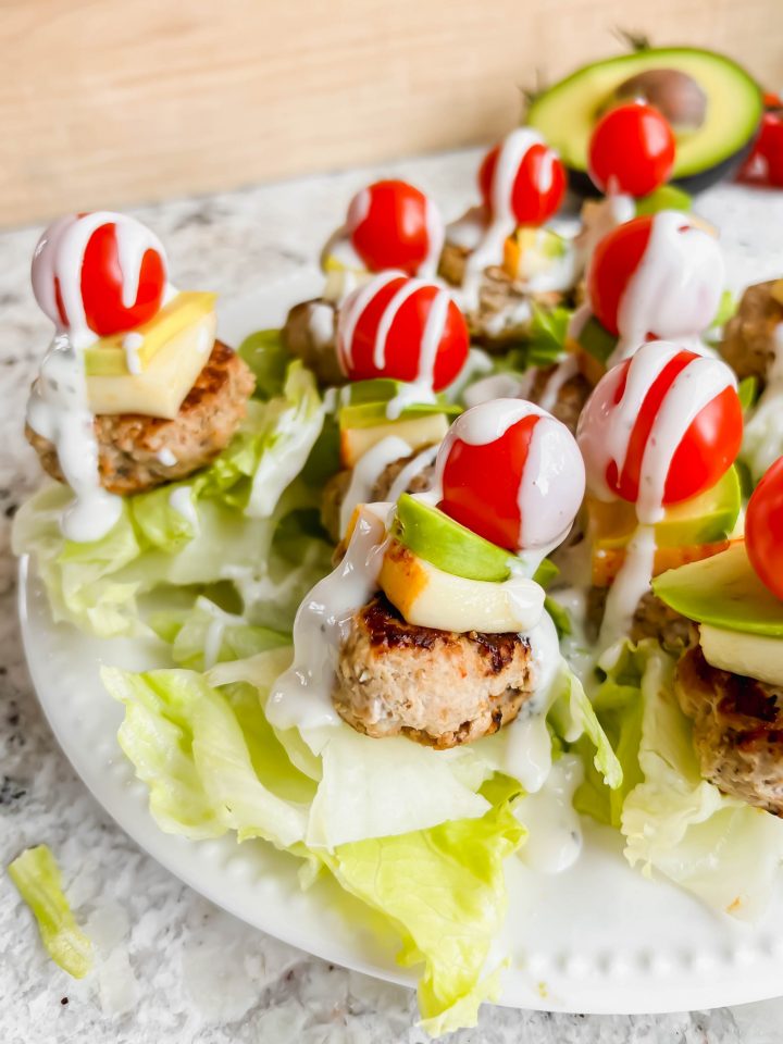 lettuce underneath a mini turkey burger on a stick topped with cheese, avocado and a cherry tomato drizzled in ranch on white plate on marble counter.