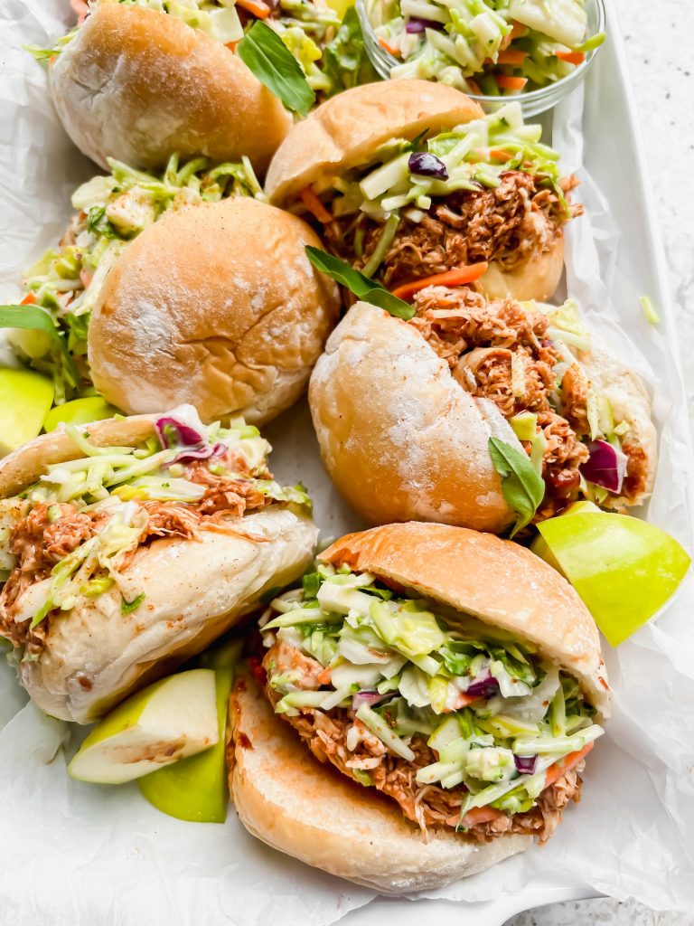 six pulled bbq chicken sliders on their side with chicken and fresh slaw pouring out on white parchment paper.