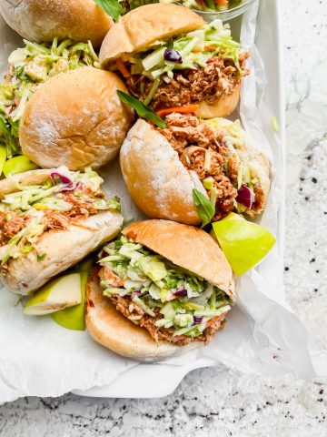 five pulled bbq chicken sliders on their side with chicken and fresh slaw pouring out on white parchment paper.