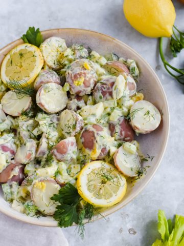 creamy healthy lemon dill potato salad mixed together garnished with lime wedges and fresh herbsin white bowl.
