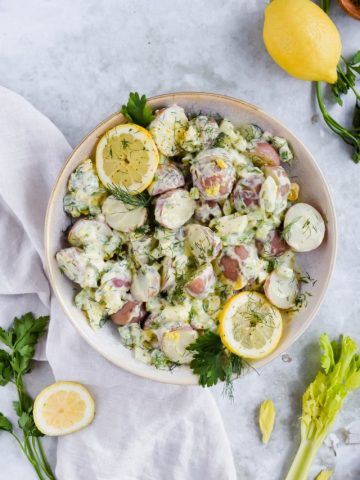 healthy lemon dill potato salad mixed together in white bowl on white background surrounded by additional recipe ingredients.