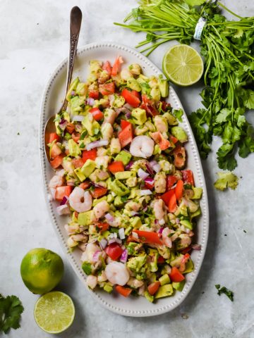 avocado shrimp ceviche salad on white serving dish beside a bunch of fresh cilantro and lime halves on gray background.