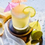 banana margarita with two drink umbrellas inserted into cocktail garnished with fresh lime.