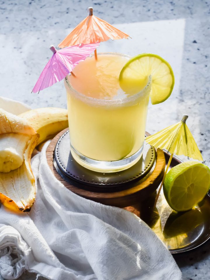 banana margarita with two drink umbrellas inserted into cocktail garnished with fresh lime.