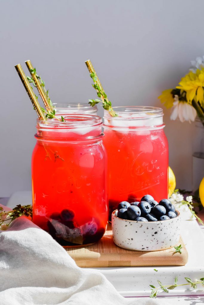 three blueberry vodka lemonades on wood board beside bowl of fresh blueberries.