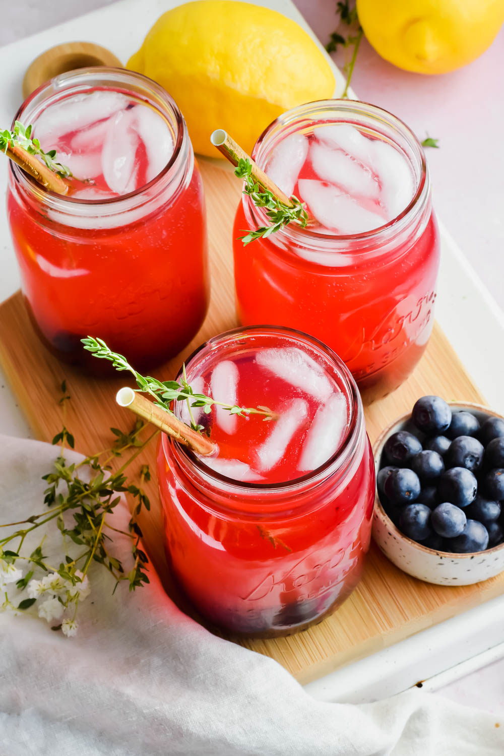 three blueberry vodka lemonades on wood board beside bowl of fresh blueberries.