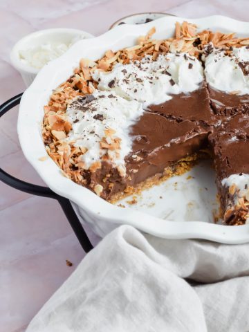 sliced chocolate coconut cream pie decorated with whipped cream and toasted coconut with one slice taken out of white pie dish.