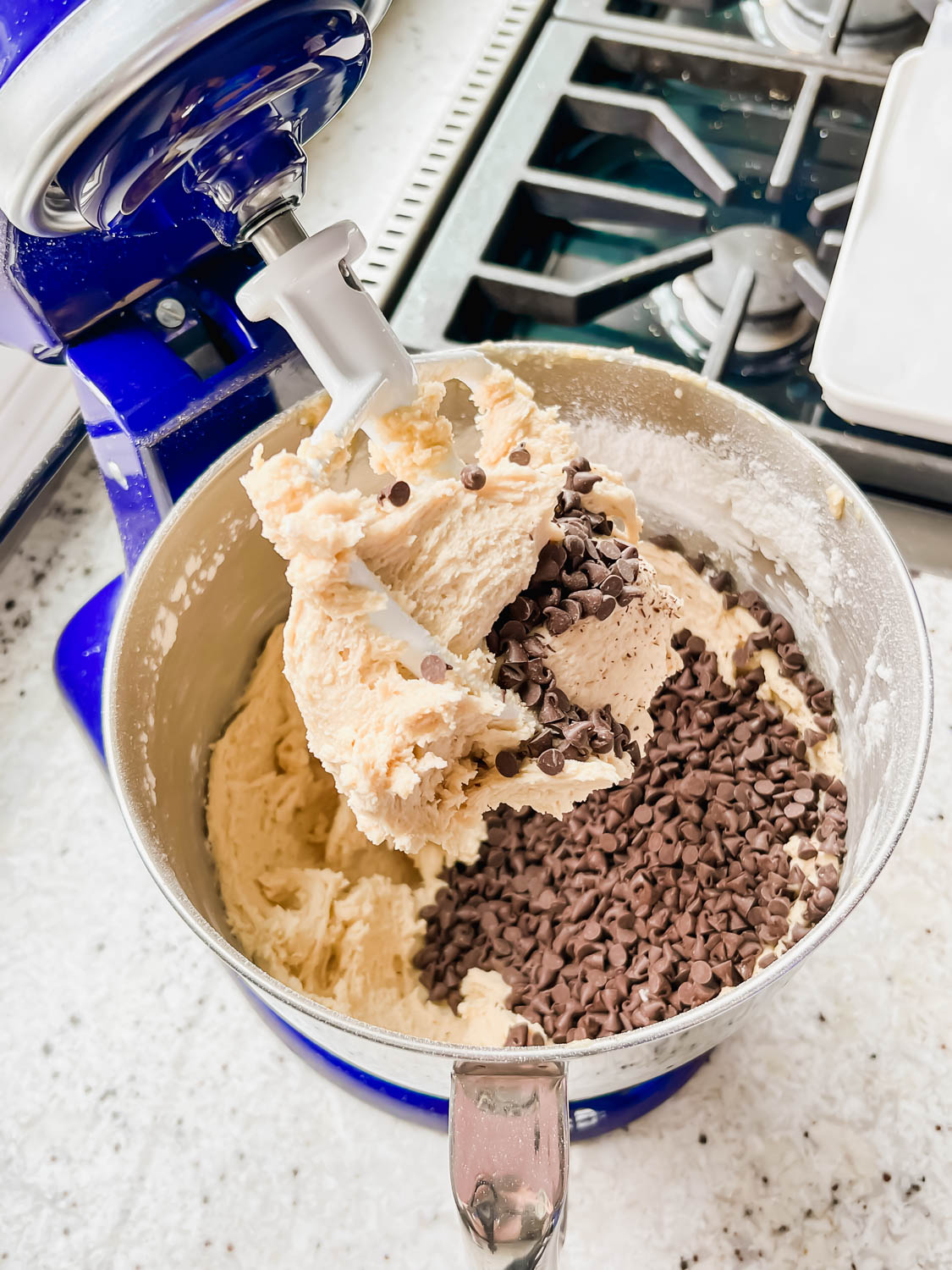 raw cookie dough in blue stand mixer with chocolate chips poured on top before being mixed in.