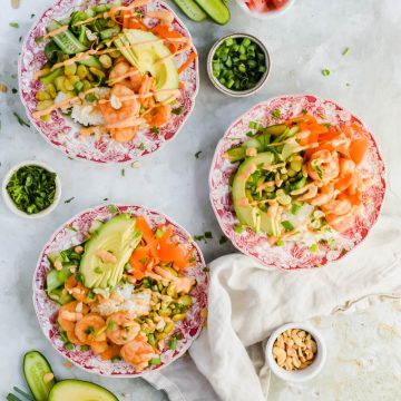 three Crunchy edamame salad bowls plated and garnished with chopped green onion and spicy mayo, surrounded by additional salad bowl ingredients.