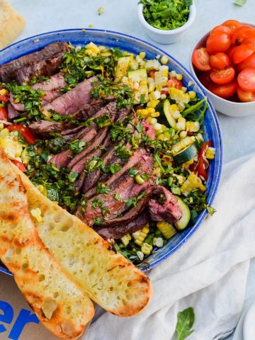 Grilled flank steak with chimichurri on top of a bed of grilled vegetables being tossed in a blue bowl with two halves of a grilled baguette on top of the salad beside a small bowl of halved cherry tomatoes and another bowl of fresh herbs.