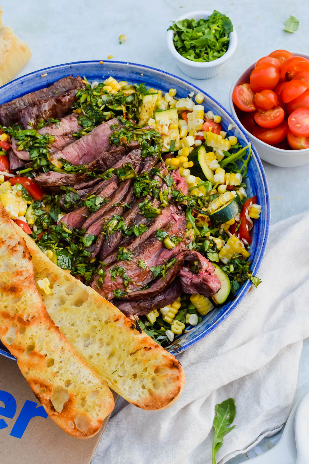 Grilled flank steak with chimichurri on top of a bed of grilled vegetables being tossed in a blue bowl with two halves of a grilled baguette on top of the salad beside a small bowl of halved cherry tomatoes and another bowl of fresh herbs.