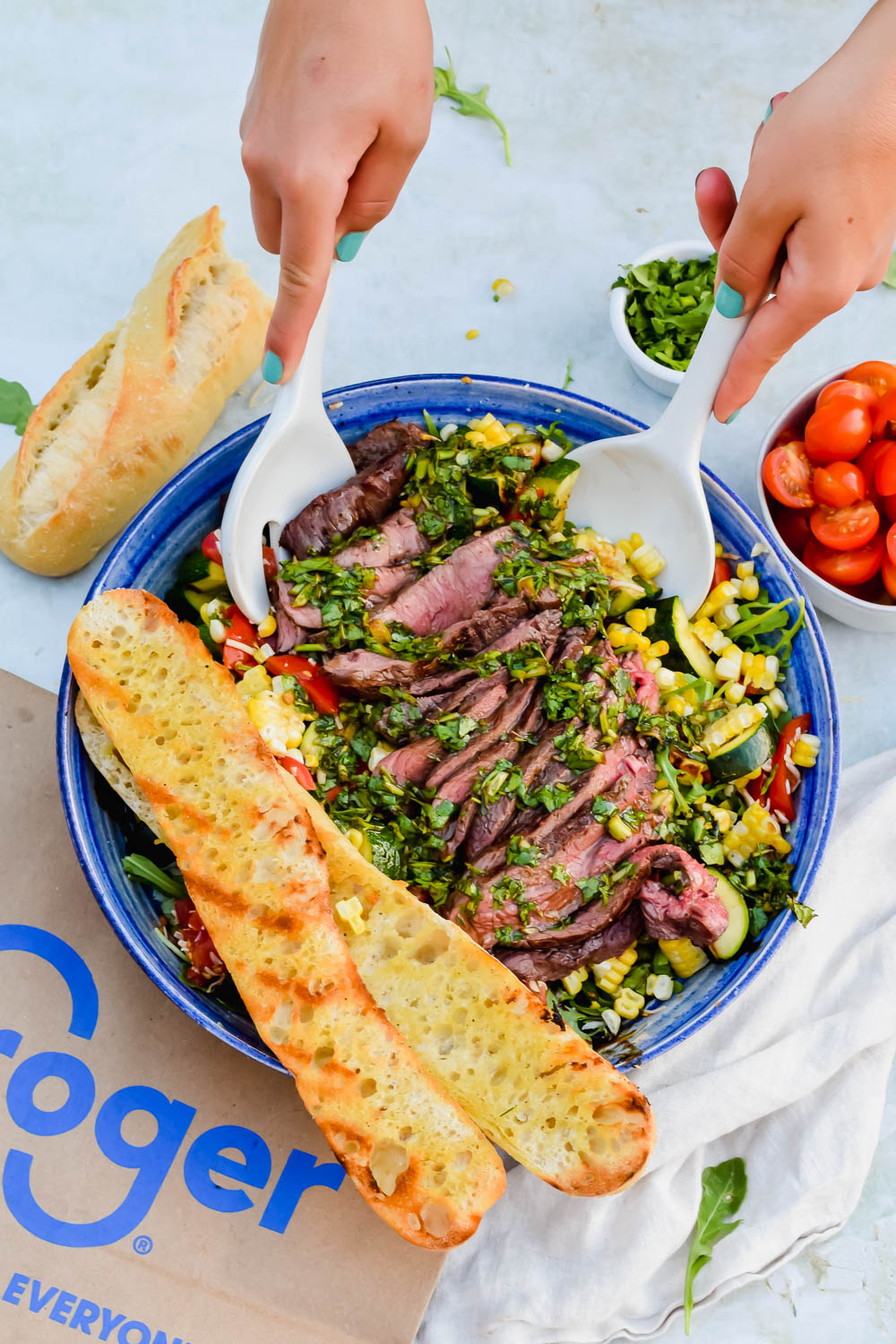 Grilled flank steak with chimichurri on top of a bed of grilled vegetables being tossed in a blue bowl.