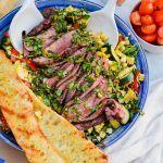 Grilled flank steak with chimichurri on top of a bed of grilled vegetables being tossed in a blue bowl.