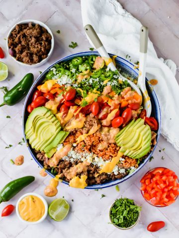 Finished healthy 7 layer salad bowl garnished with queso and avocado slices in large bowl with smaller bowl of additional ingredients surrounding.