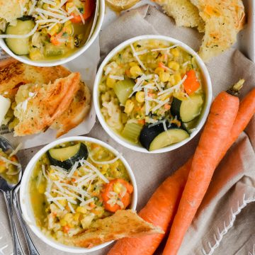 three small white ramekins filled with Healthy Summer Zucchini Corn Chowder garnished with parmesan beside toasted bread, fresh carrots, and silver spoons.