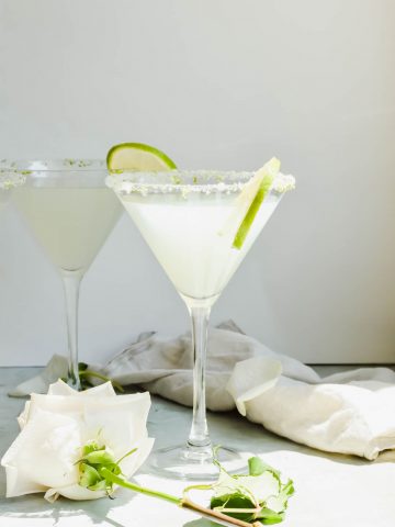 two lemon drop martinis garnished with lime slices on gray background beside white flower and white dish towel.