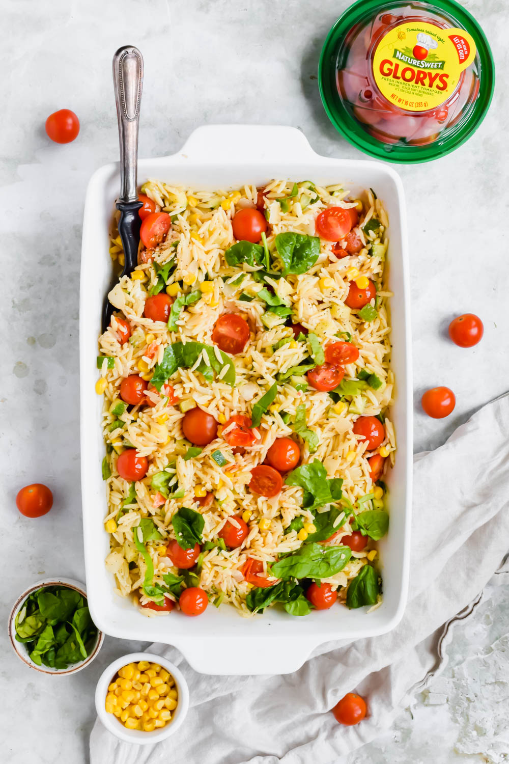 prepared summer orzo salad in white baking dish surrounded by additional recipe ingredients on white background.