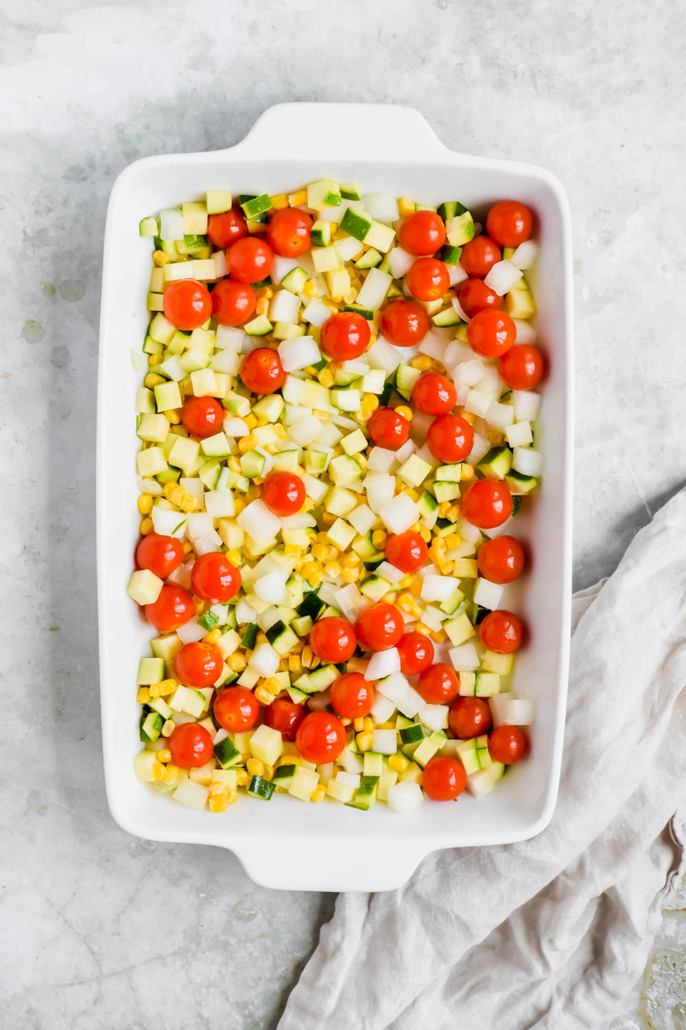 uncooked chopped zucchini, onion, corn, and baby tomatoes in white baking dish on white background.