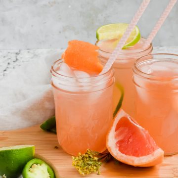 three spicy mezcal palomas in glass jars with striped straw on wood cutting board surrounded by grapefruit slices, lime wedges, and jalapeños.