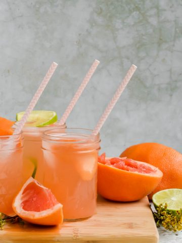 three spicy mezcal palomas in glass jars with striped straw on wood cutting board surrounded by grapefruit slices with gray background.
