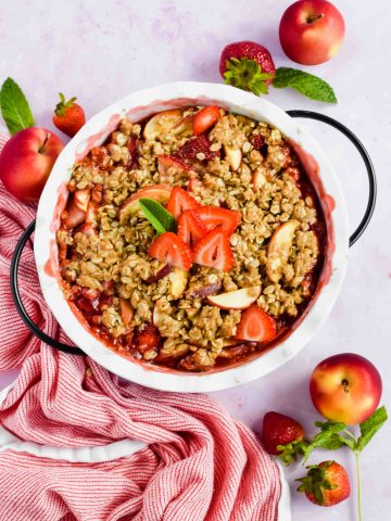 baked strawberry apple crisp in white pie dish toped with fresh strawberry slices next to red and white striped tea towel.