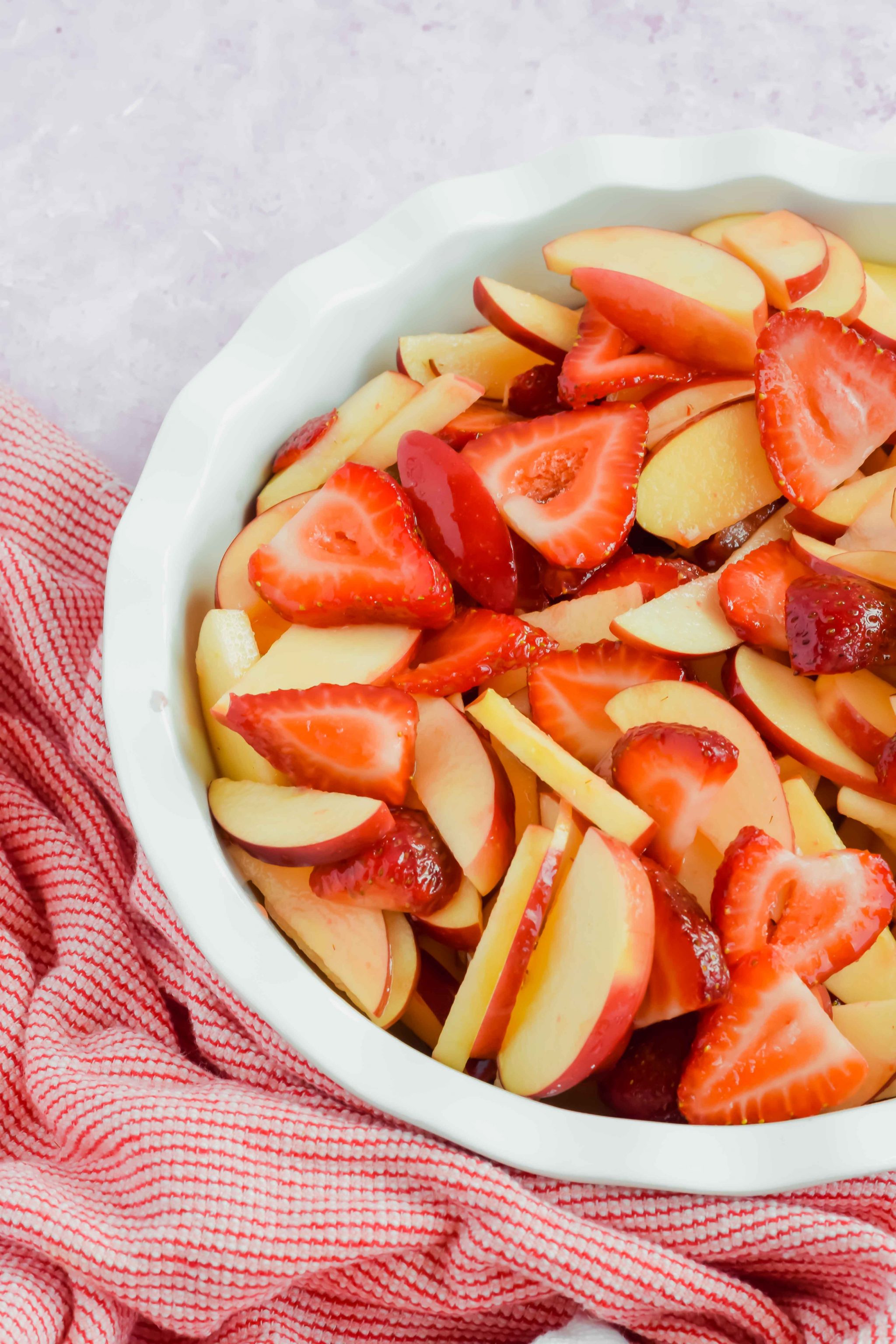 sliced apples and strawberries in white pie dish beside red and white striped dish towel.