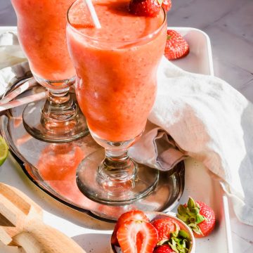 two strawberry mango daiquiris in glasses garnished with a fresh strawberry and white straws on silver tray.