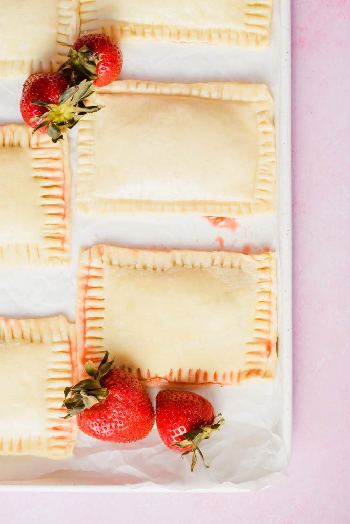 Unbaked strawberry pop tarts beside fresh strawberries on white parchment on pink background.